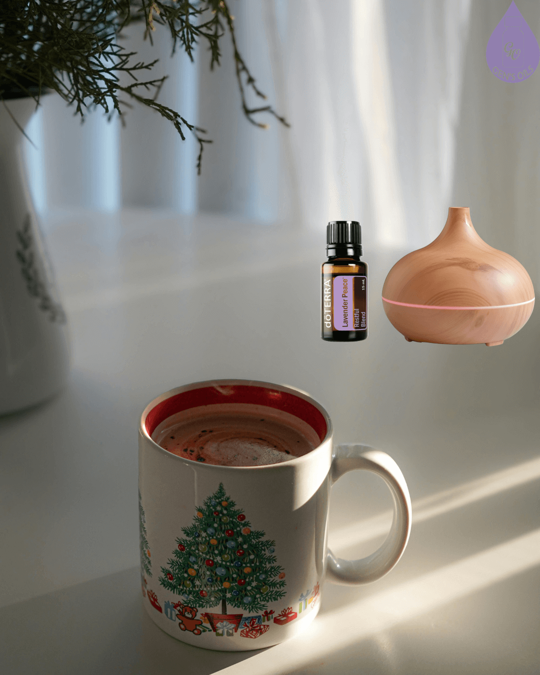 A mug with a Christmas tree design sits on a white table next to greenery in a vase, with a diffuser & bottle of Lavender Peace essential oil.