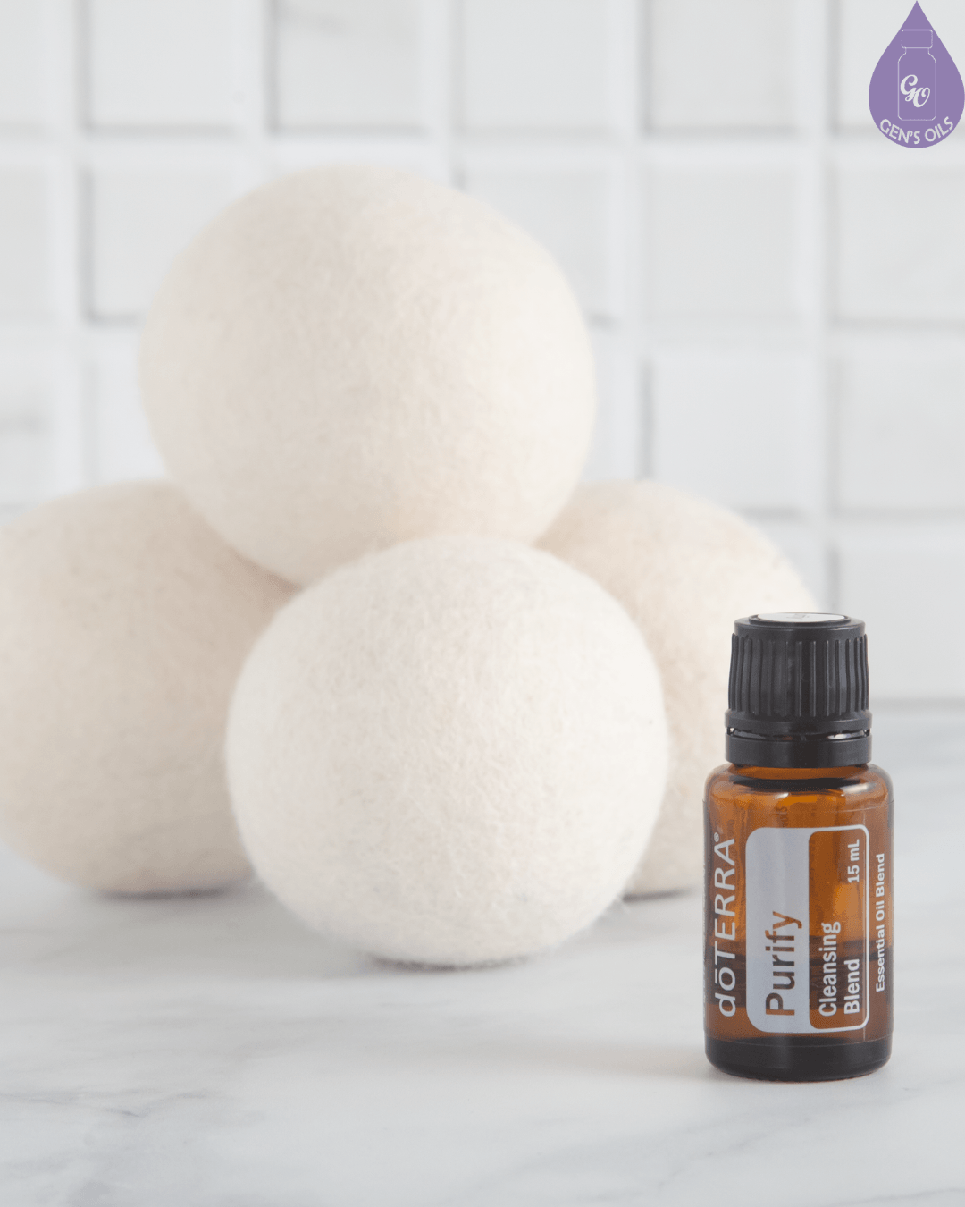 A small brown bottle of doTERRA Purify Cleansing Blend essential oil sits in front of a pile of four white wool dryer balls against a white tiled background.