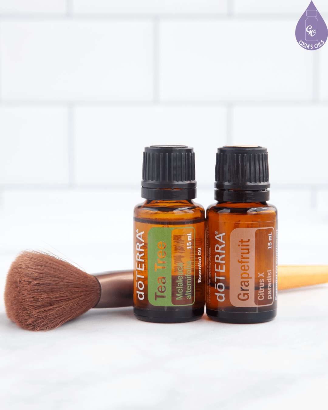 Two doTERRA essential oil bottles, one Tea Tree and one Grapefruit, sit beside a makeup brush on a marble surface.