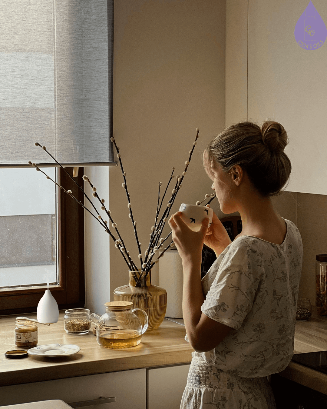 A person sips from a mug in a kitchen with a clear teapot and a vase of pussy willows & a diffuser going in the background.