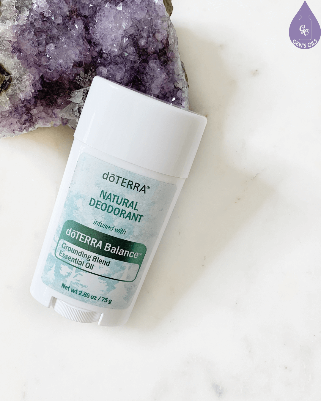 A white stick of dōTERRA Natural Deodorant infused with dōTERRA Balance Grounding Blend Essential Oil sits next to a large purple amethyst geode on a white marble surface.
