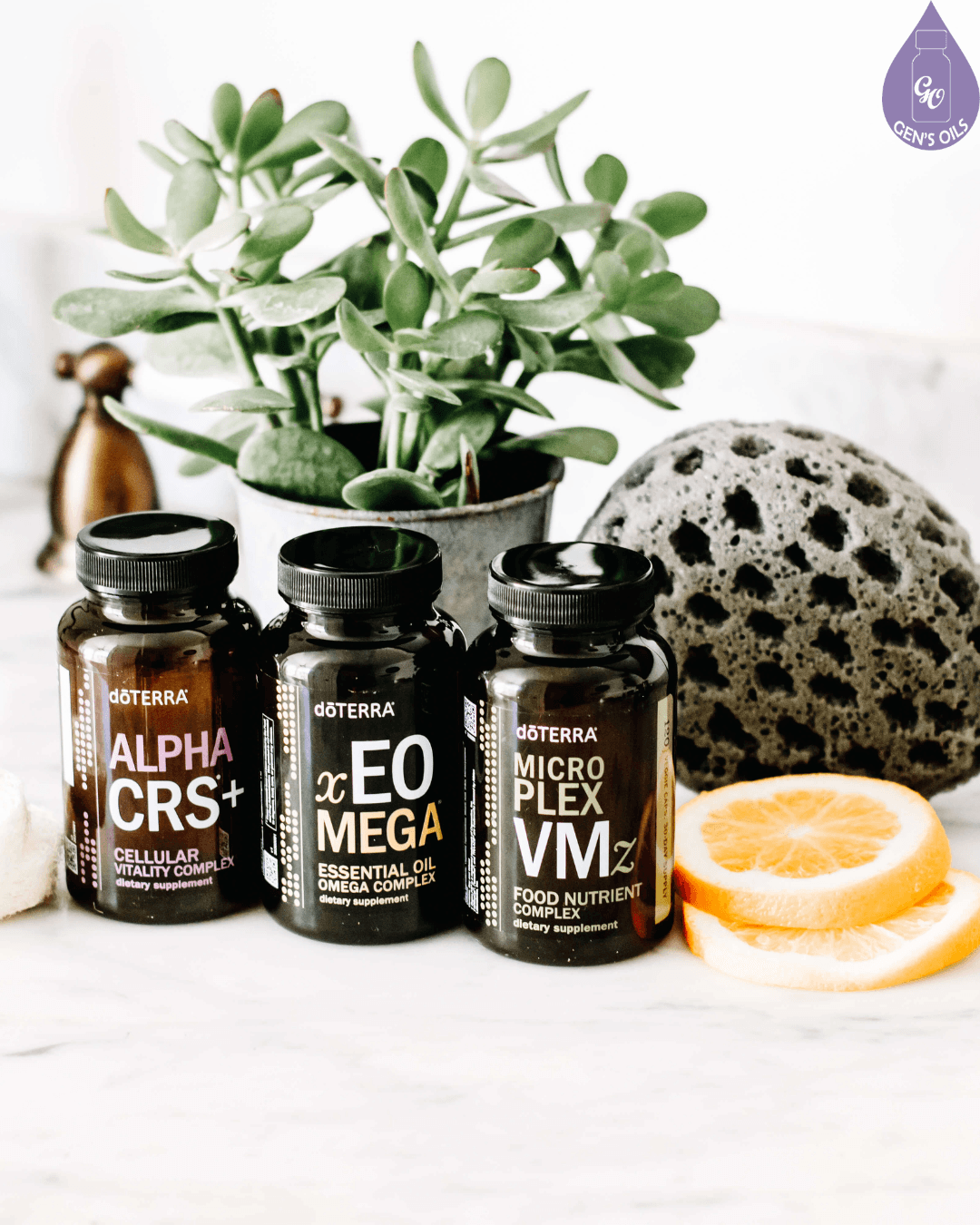 Three bottles of DoTERRA supplements, Alpha CRS+, EO Mega, and Micro Plex VMz, are displayed on a marble countertop next to a potted plant, a decorative sponge, and orange slices.