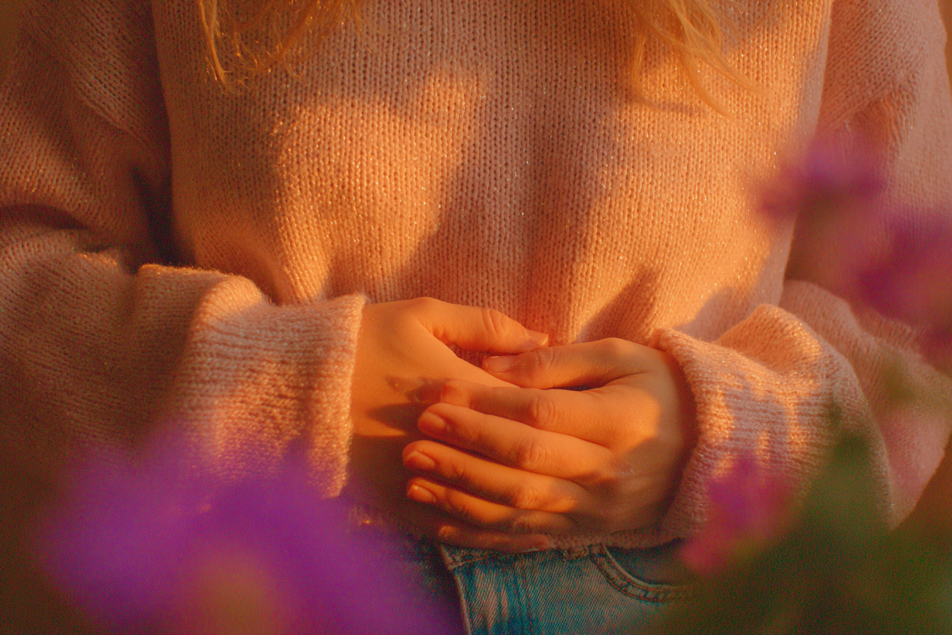 A woman gently holding her hands over her lower belly during the two week wait, soft afternoon light, warm tones of peach and dusty purple